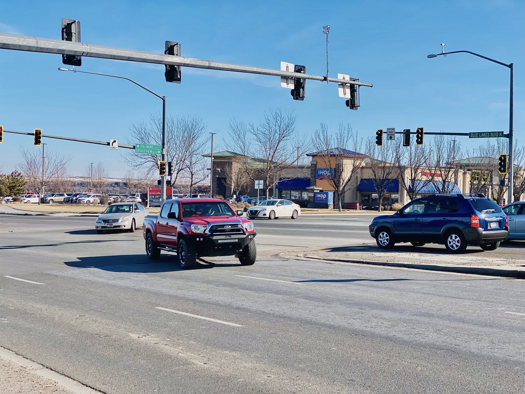 Image of a busy intersection at Blue Lakes Boulevard and Fillmore Street with vehicles, clear sky, trees, structures, and traffic signals.